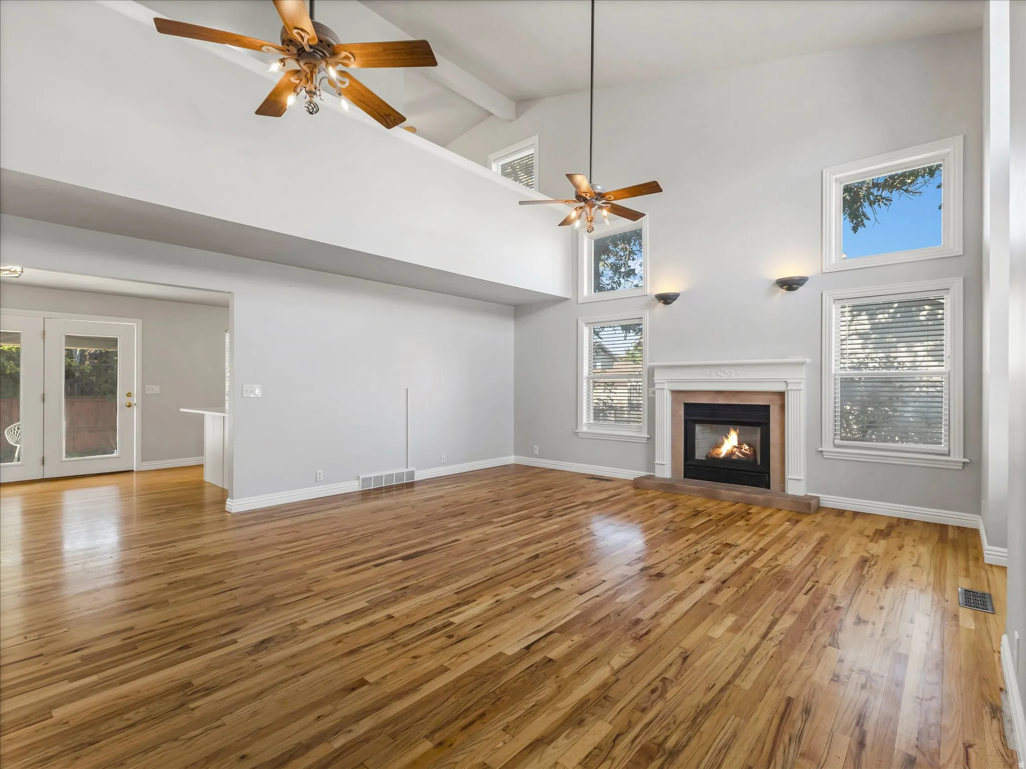 Spacious living area featuring high vaulted ceilings, natural wood-finish flooring, a fireplace with a white mantel, multiple windows for abundant illumination, and dual ceiling fans