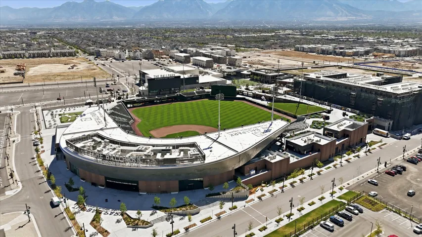 Aerial perspective showcasing a modern sports stadium with a manicured green field, surrounded by developed urban infrastructure and distant mountains