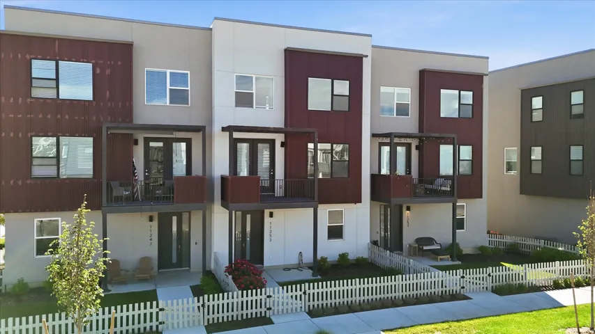 Modern townhome exteriors featuring white and maroon vertical siding, private balconies with dark railings, and a contrasting white picket fence enclosing a landscaped front yard