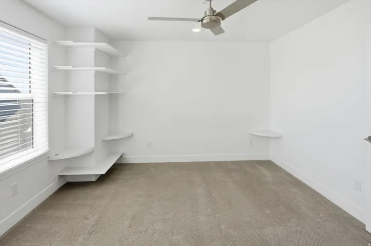 Room featuring a built-in shelving unit, neutral carpet, a ceiling fan with integrated lighting, and a window with blinds