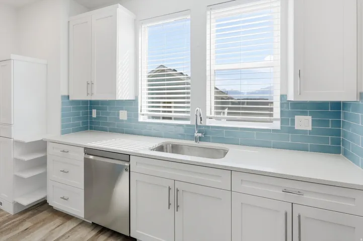 Kitchen featuring white shaker cabinetry, an integrated stainless steel dishwasher, a single-basin sink with a gooseneck faucet, light-toned countertops, and a blue subway tile backsplash
