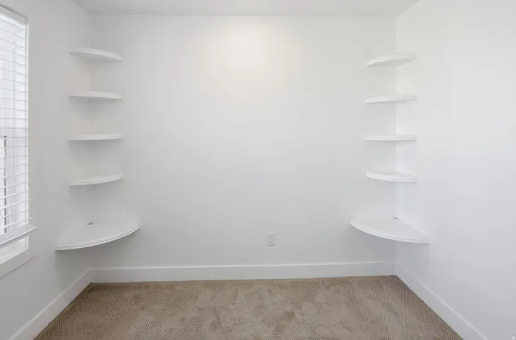 Room featuring dual built-in corner shelving units, plush carpet flooring, white painted walls, and a window with horizontal blinds