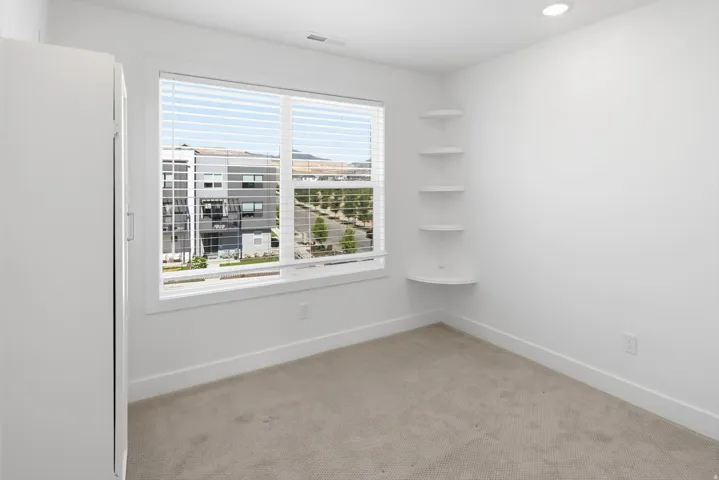 Bright room featuring a large window with blinds, recessed lighting, built-in corner shelving, and light-toned carpeting