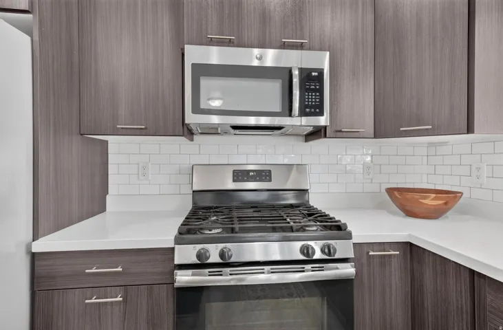 Modern kitchen featuring a stainless steel gas range, over-the-range microwave, white subway tile backsplash, white countertops, and wood-finish cabinetry with bar pull hardware