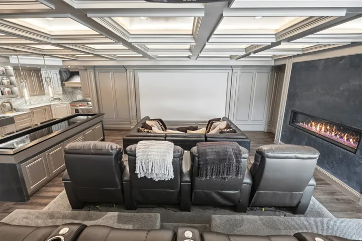 Entertainment room featuring a coffered ceiling with integrated lighting, a built-in linear fireplace with a dark-toned accent wall, a large projection screen, and a wet bar with cabinetry and a light-toned backsplash