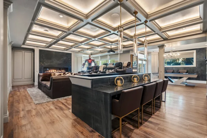 Coffered ceiling with integrated lighting overhead, featuring three pendant lights above a dark stone-top bar with seating