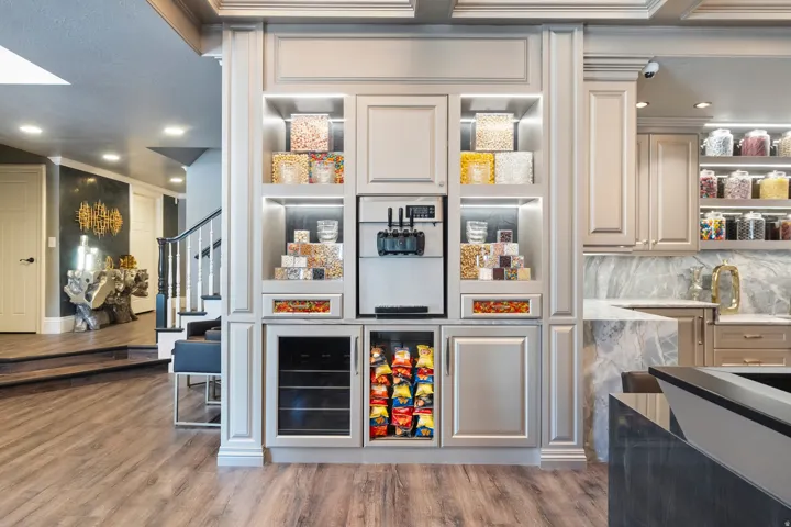 Integrated snack and beverage station featuring light-toned cabinetry with crown molding, a built-in soft-serve machine, illuminated shelving, and under-counter refrigeration units