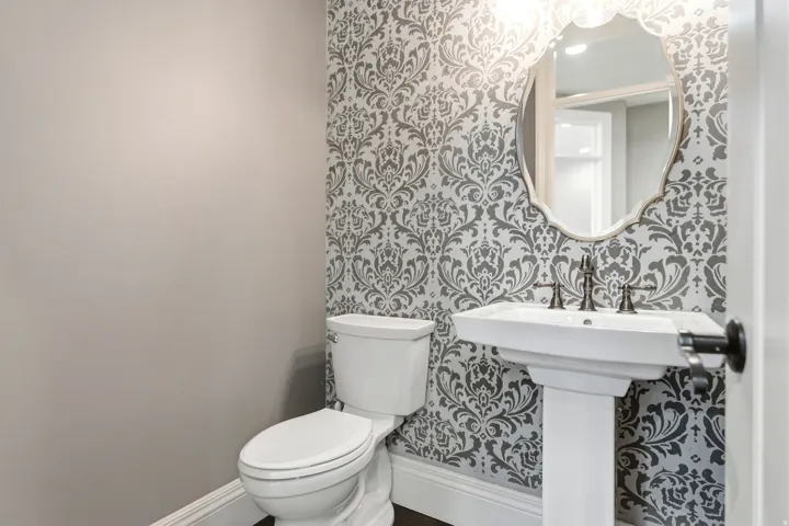 Powder room featuring a pedestal sink with dark bronze hardware, decorative patterned wallpaper, and a framed mirror with an ornate edge