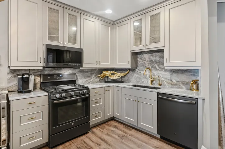 Modern kitchen featuring light-toned cabinetry, a gas range with an overhead microwave, a built-in dishwasher, a single-basin sink with a gold-tone faucet, and stone countertops paired with a matching backsplash