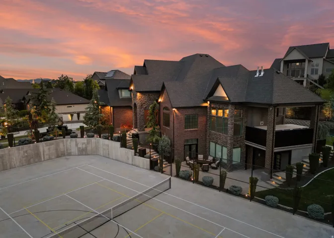 Spacious brick and stone residence featuring an outdoor sports court with netting, tiered landscaping, and an outdoor patio with seating