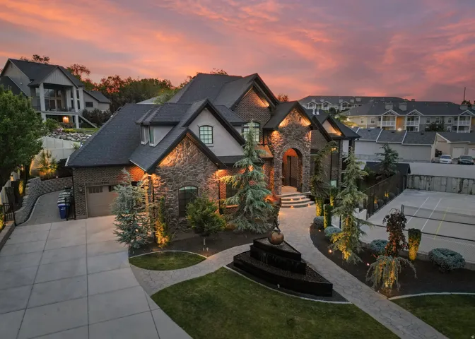 Grand stone and brick exterior featuring extensive architectural lighting, a multi-tiered water feature, manicured landscaping, a paved driveway, and a private sports court