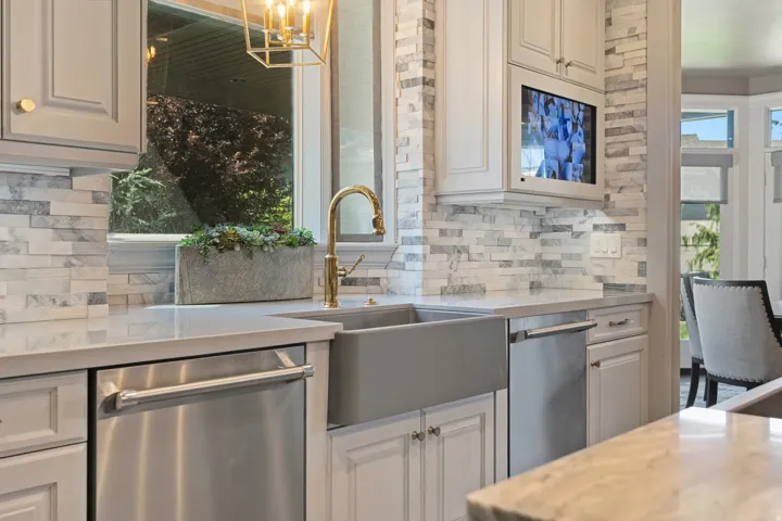 Kitchen workspace featuring a farmhouse sink with a gold-tone faucet, light-toned cabinetry, stainless steel appliances, stacked stone backsplash, and solid surface countertops
