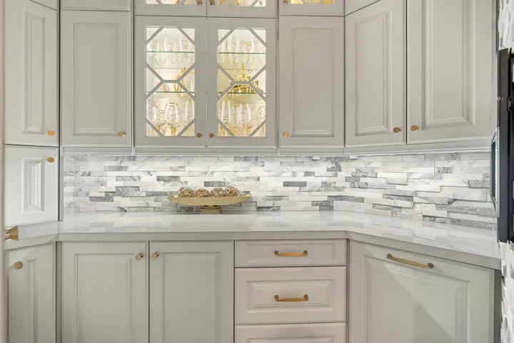 White shaker-style cabinetry with gold-toned hardware, under-cabinet lighting, glass-front display cabinets, and a multi-toned stacked stone backsplash
