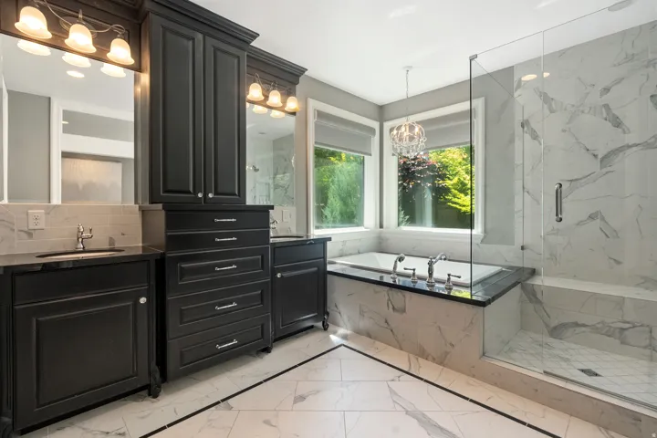 Bathroom featuring an integrated soaking tub, a frameless glass shower enclosure, a dual vanity with dark cabinetry, stone countertops, and marble-look tile flooring