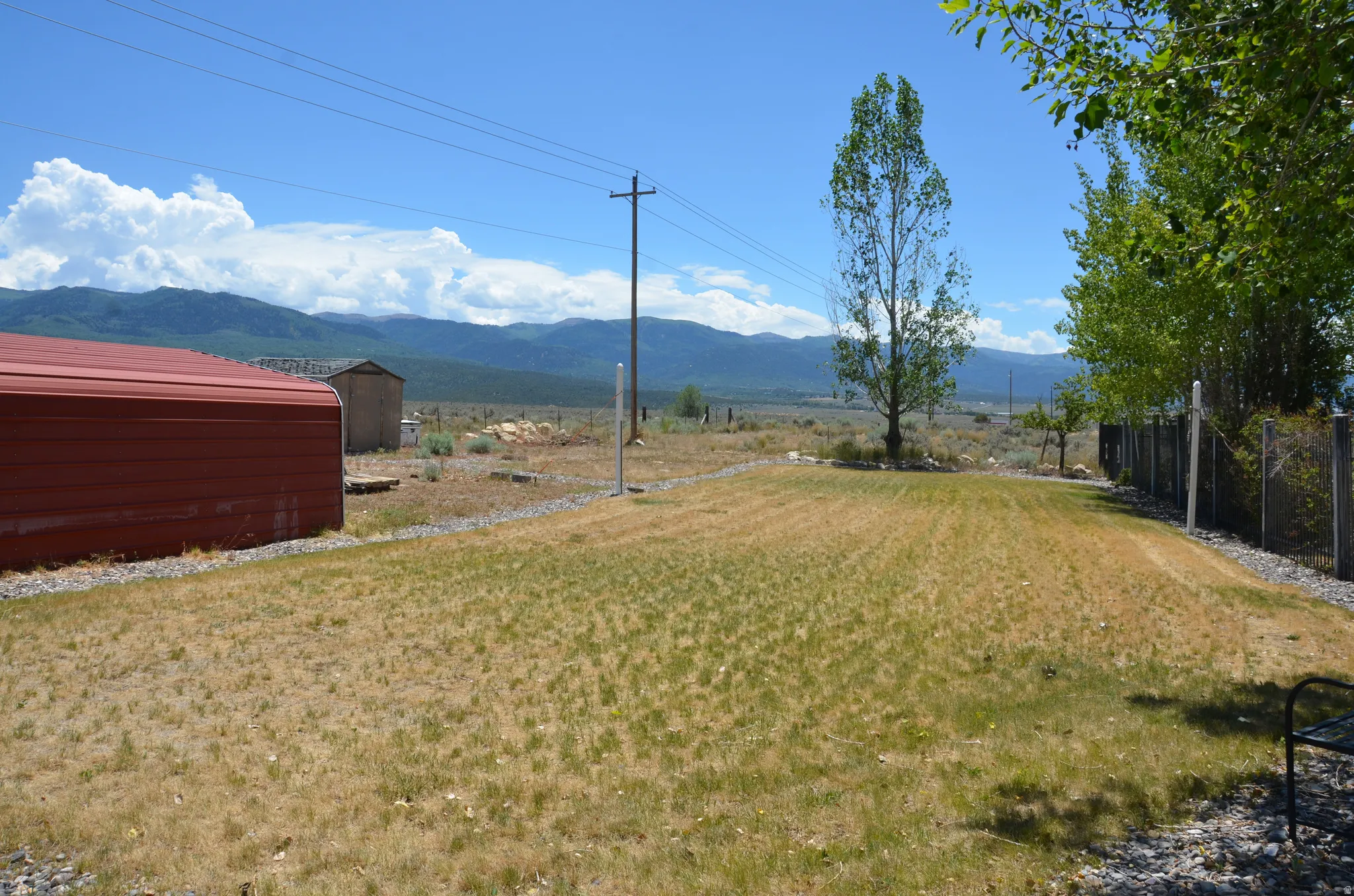 Expansive outdoor area featuring a volleyball court, two sheds, a fenced perimeter, and distant mountain scenery under a clear sky