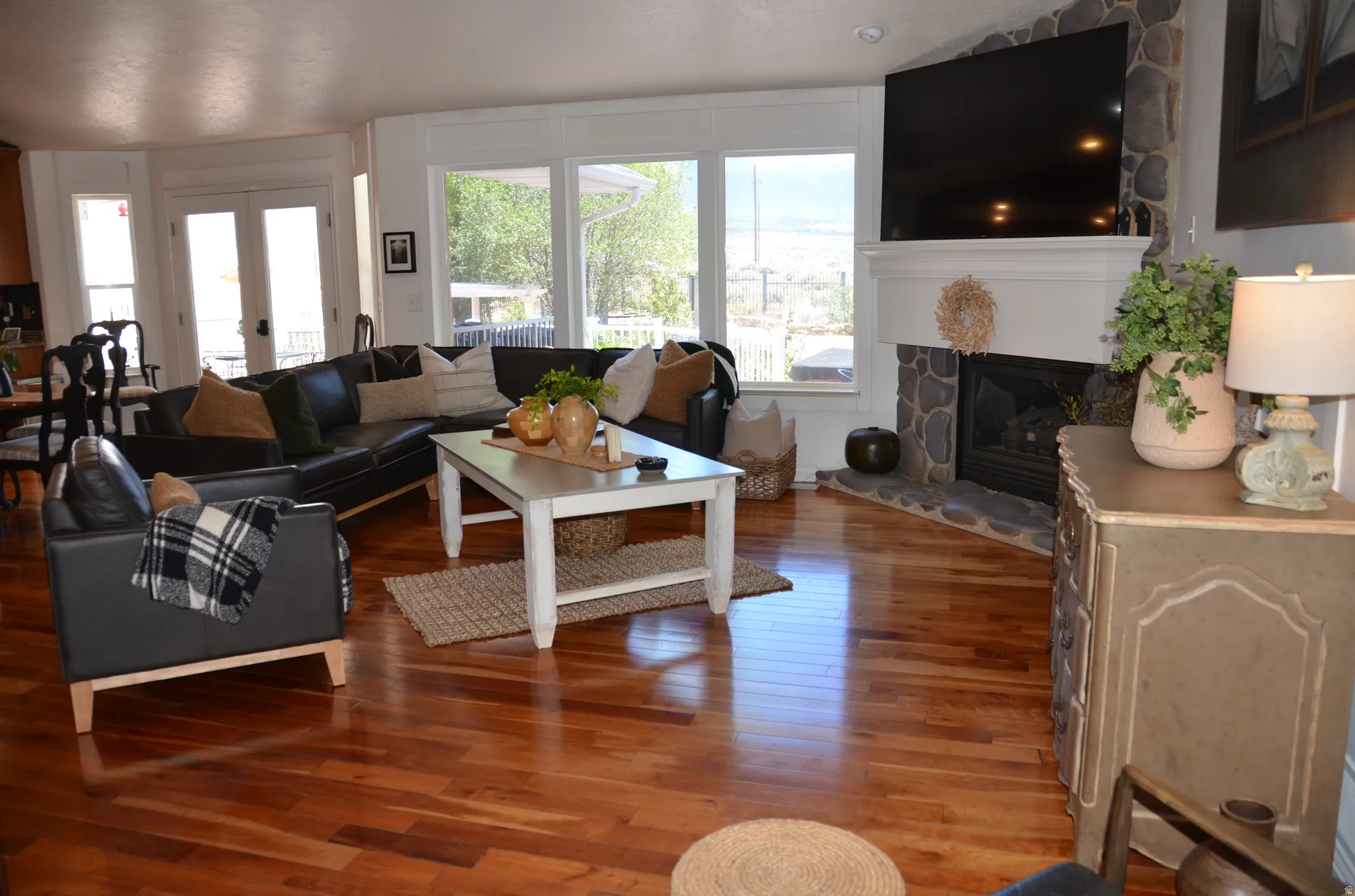 Spacious living area featuring rich wood-finish flooring, a stone-accented corner fireplace with a white mantel, and large picture windows offering abundant natural light