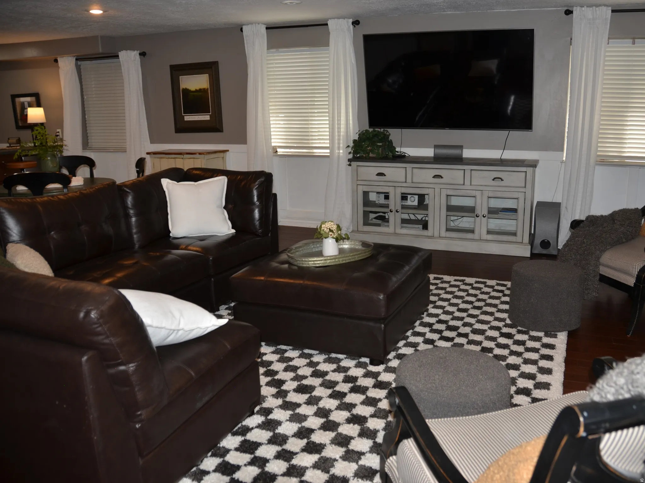 Spacious living area featuring dark wood-finish flooring, white window treatments.