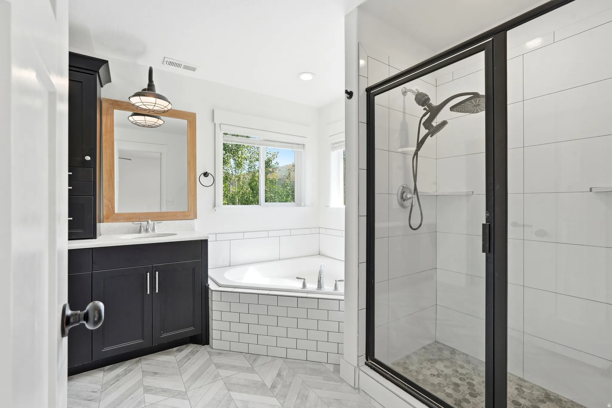 Bathroom featuring a corner soaking tub, a stand-up shower with a glass enclosure, a single vanity with dark cabinetry and a light-colored countertop, a wood-framed mirror, and patterned tile flooring