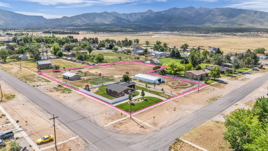 Expansive corner lot with mountain backdrop, featuring a single-story residence with a brown roof, multiple outbuildings, mature trees, and a white perimeter fence