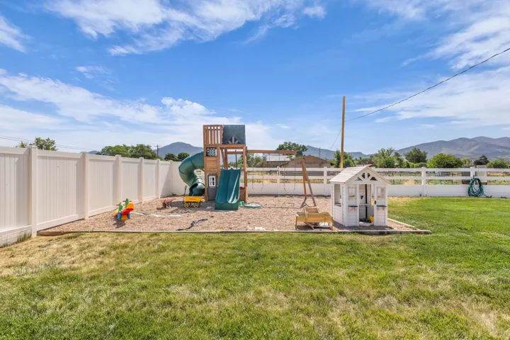 Expansive backyard featuring a dedicated play area with a wooden playset, slide, and tube slide, a swing set, and a white playhouse, all set on a gravel base