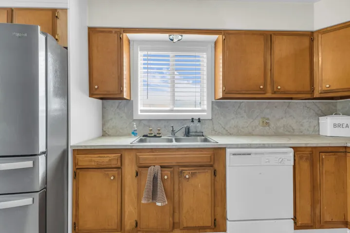 Kitchen area featuring wood cabinetry, a double basin stainless steel sink, a window with blinds, a built-in dishwasher, and a stainless steel refrigerator