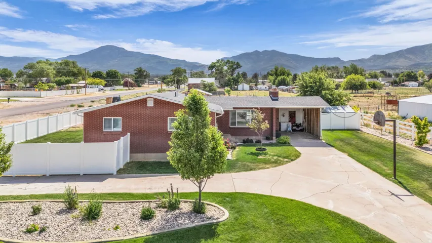Red brick ranch-style residence featuring a lush green lawn, mature trees, a paved driveway, and white perimeter fencing