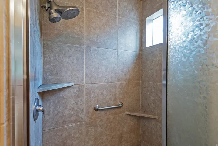 Tiled shower with a dual shower head, built-in corner shelving, integrated grab bar, and translucent window