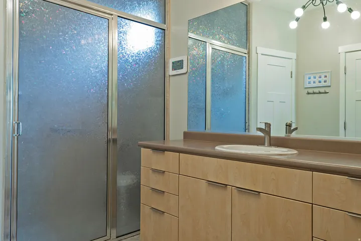 Bathroom featuring a glass-enclosed shower with textured privacy glass, a spacious vanity with light wood-finish cabinetry, a solid surface countertop, an undermount sink, and a contemporary faucet