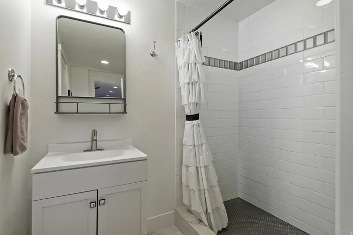 Compact bathroom featuring a shower with white subway tile and a decorative tile accent band, a small-scale white vanity with an integrated sink, a brushed nickel faucet, and a wall-mounted mirror with a storage shelf