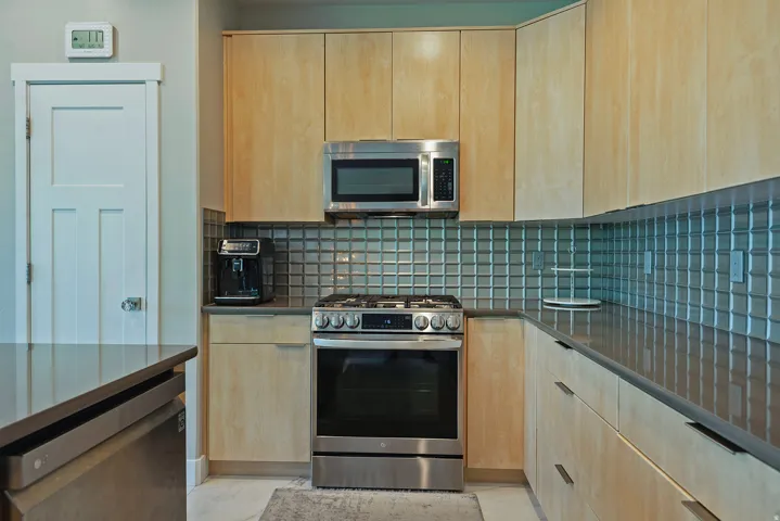 Modern kitchen featuring light wood-finish cabinetry, stainless steel appliances, a glass tile backsplash, and dark countertops