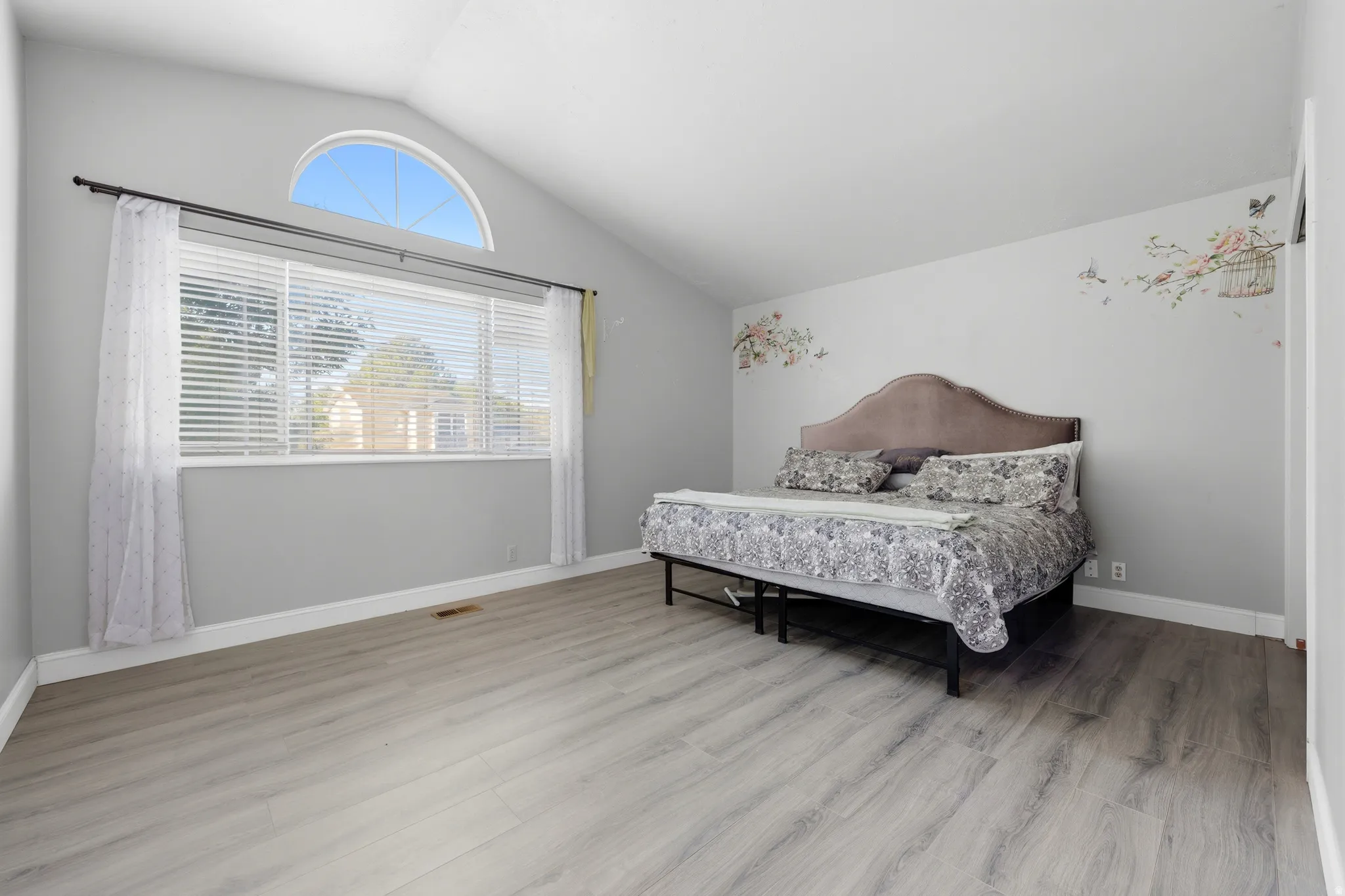 Vaulted ceiling bedroom featuring wood-finish flooring, a large horizontal window with blinds, and an arched transom window