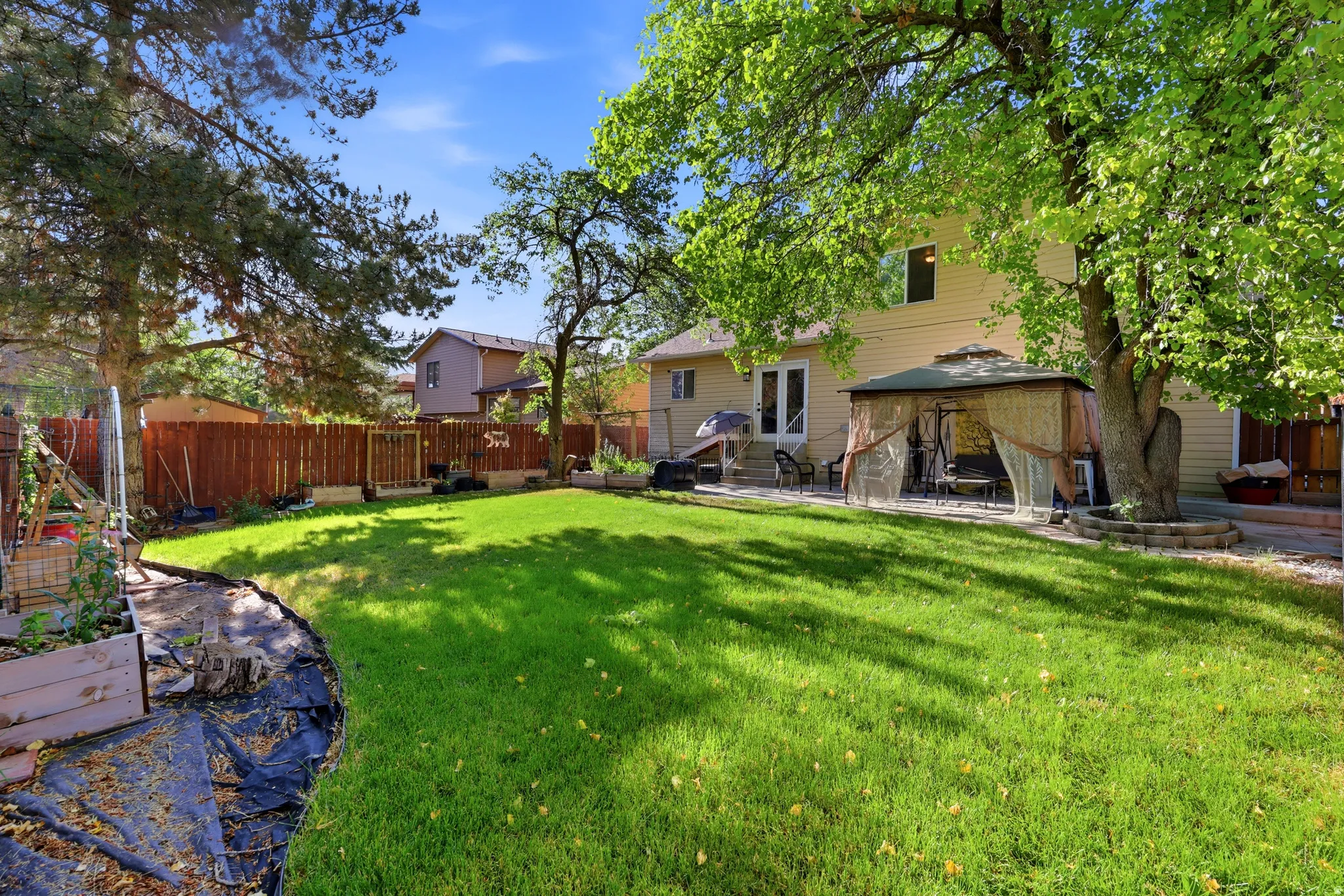Expansive backyard featuring a manicured lawn, established shade trees, a patio area, and raised garden beds
