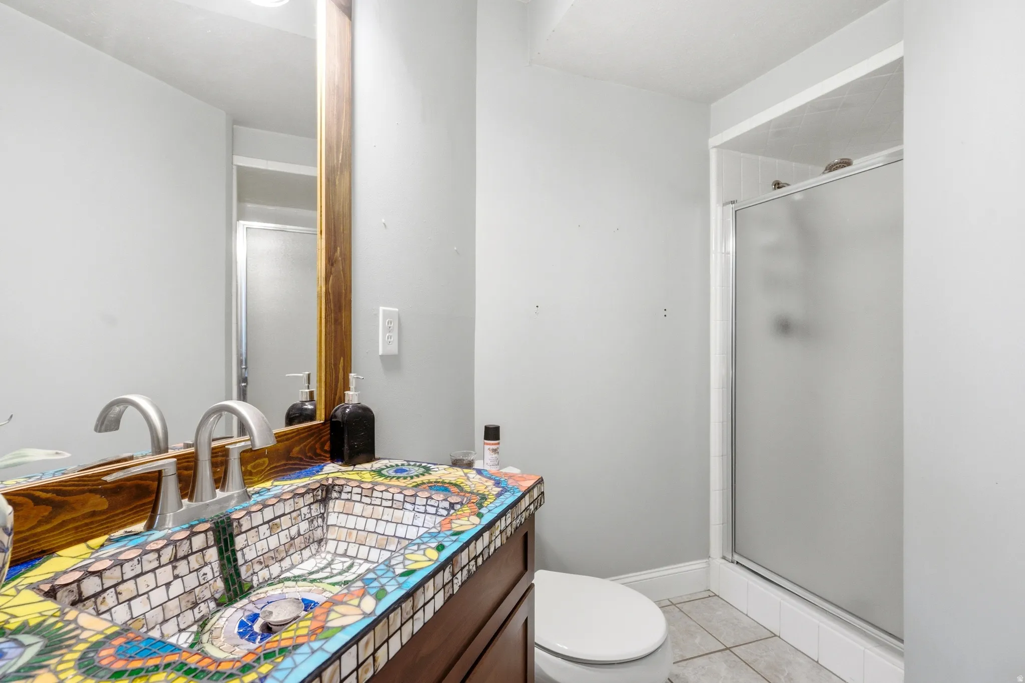 Bathroom featuring a mosaic-tiled vanity and integrated sink, a large mirror with a wood-finish frame, and a glass-enclosed shower with a frosted panel