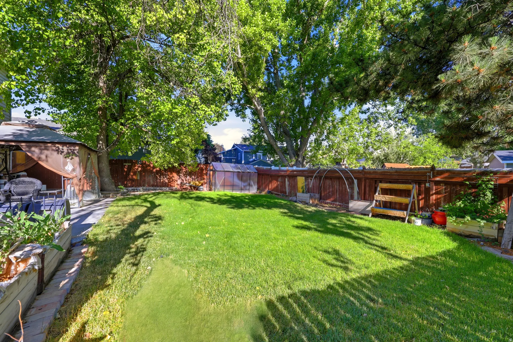 Expansive rear yard featuring a lush lawn, mature trees, wood fencing, and a dedicated garden area with a greenhouse structure and raised garden beds