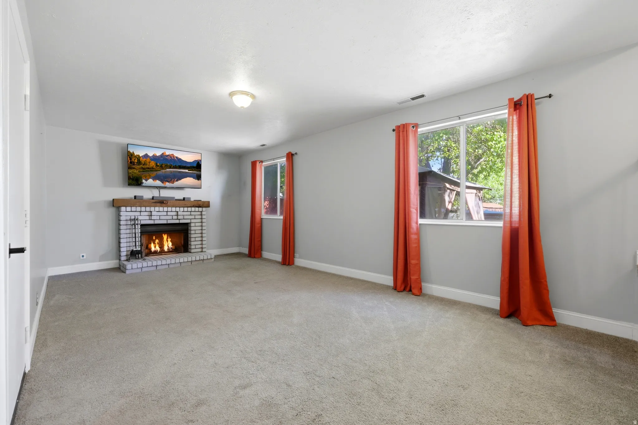 Spacious room featuring a white-painted brick fireplace with a wood mantel, light gray walls, and carpet flooring