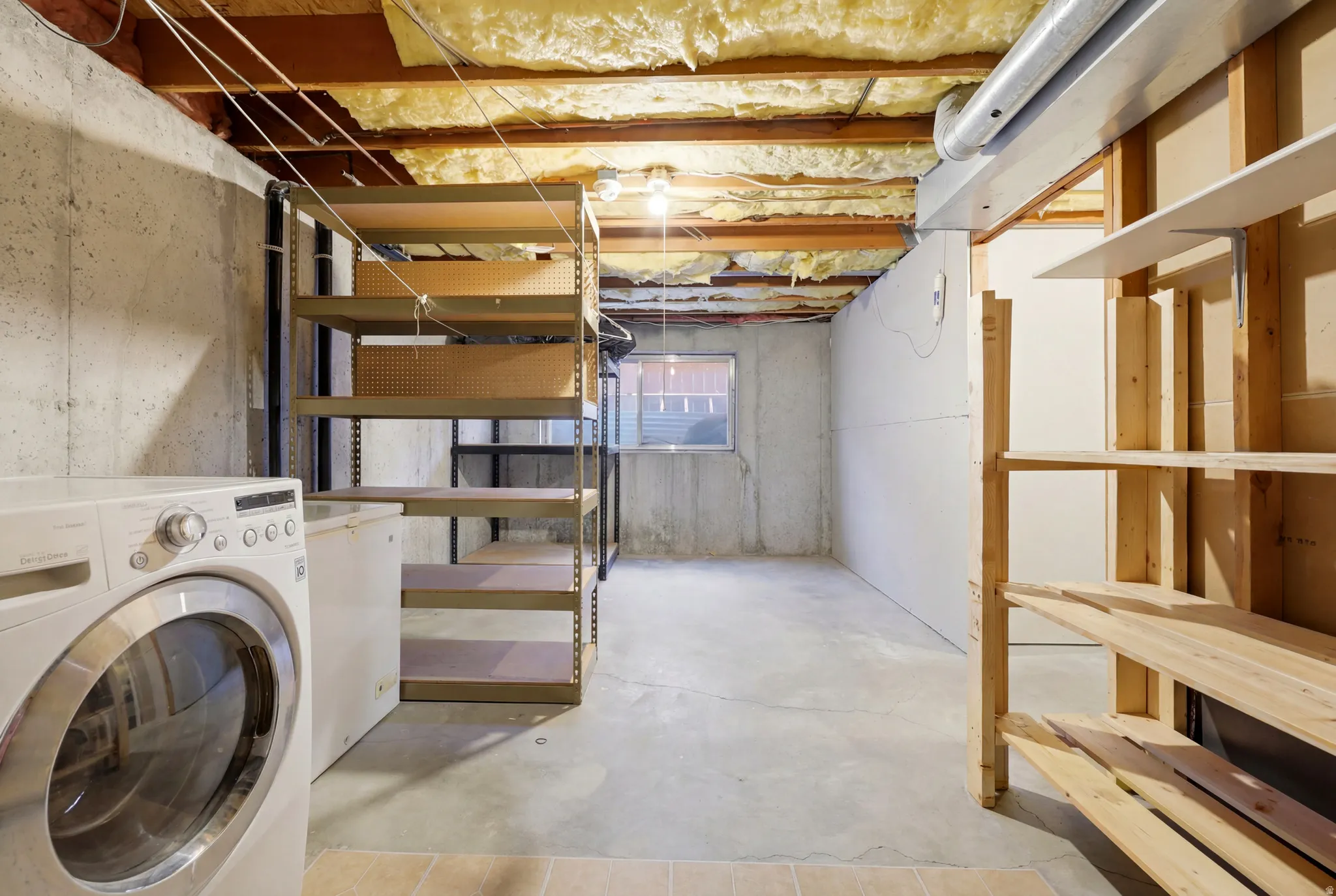 Unfinished basement featuring exposed joists, concrete walls, a small window, built-in shelving, and a laundry area