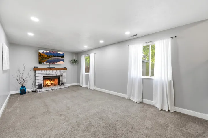Recessed lighting, brick fireplace with wood mantel, two double-hung windows, and grey carpeting