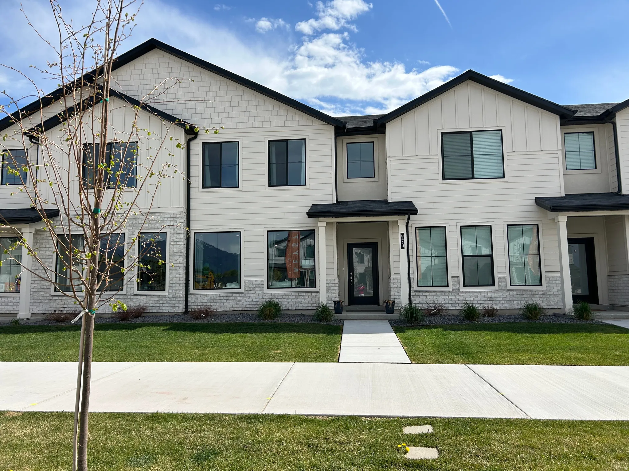 Contemporary townhome exterior featuring light-toned siding, black-framed windows, dark rooflines, stone-look accents, and a concrete walkway