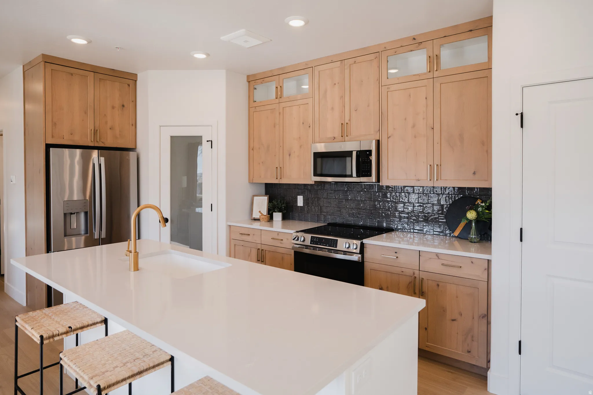 Kitchen featuring a large central island with an undermount sink and golden faucet, natural wood cabinetry, stainless steel appliances, and a dark subway tile backsplash
