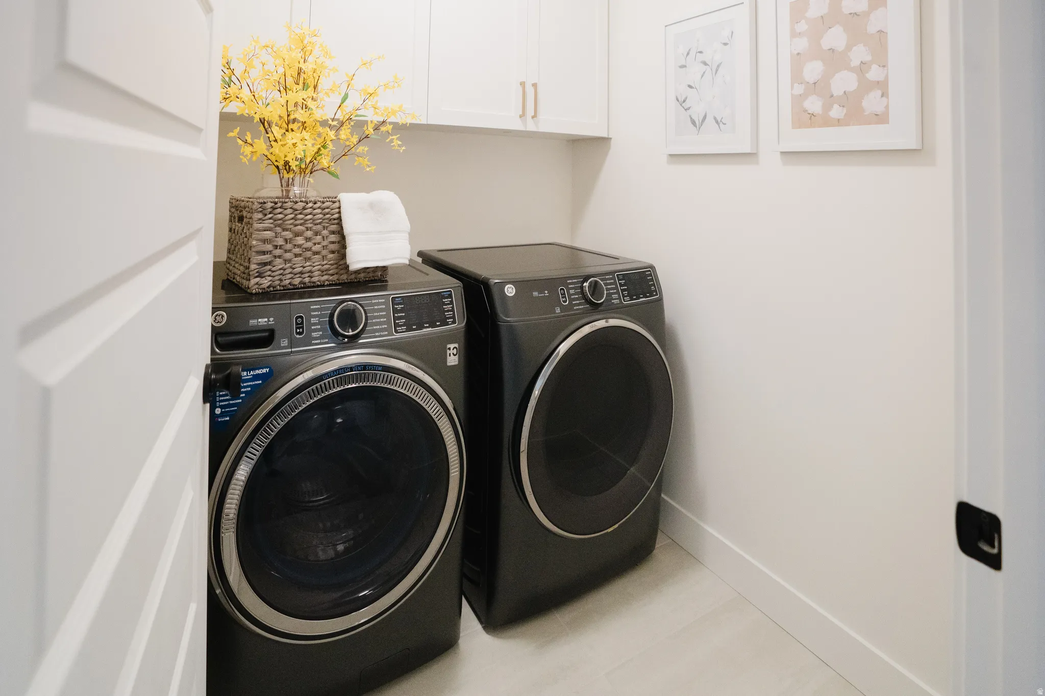 Dedicated laundry area featuring a front-loading washer and dryer, white shaker-style cabinetry with gold-tone hardware, and light-toned flooring