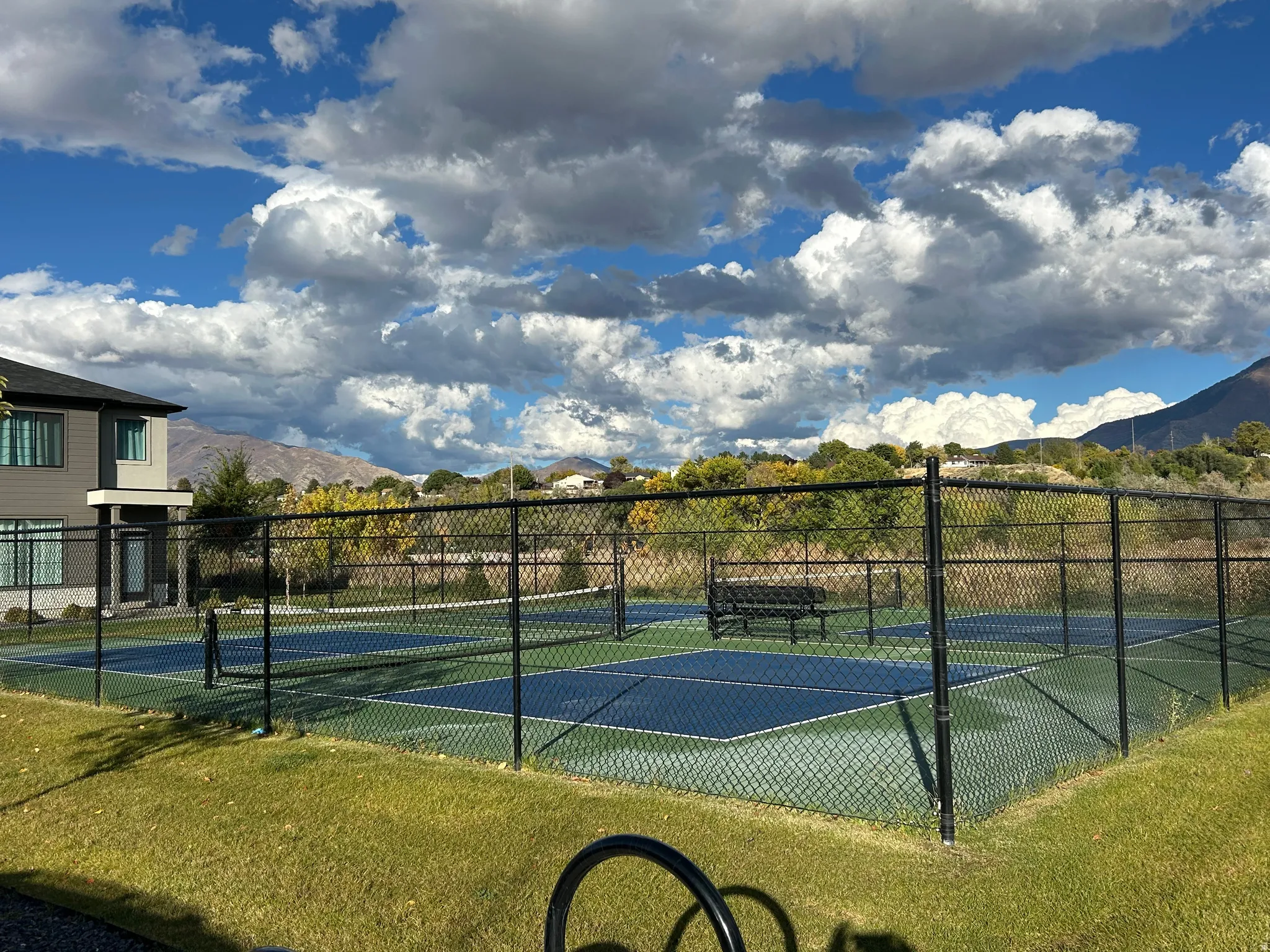Fenced outdoor court with blue and green surfacing, surrounded by a manicured lawn and distant mountains