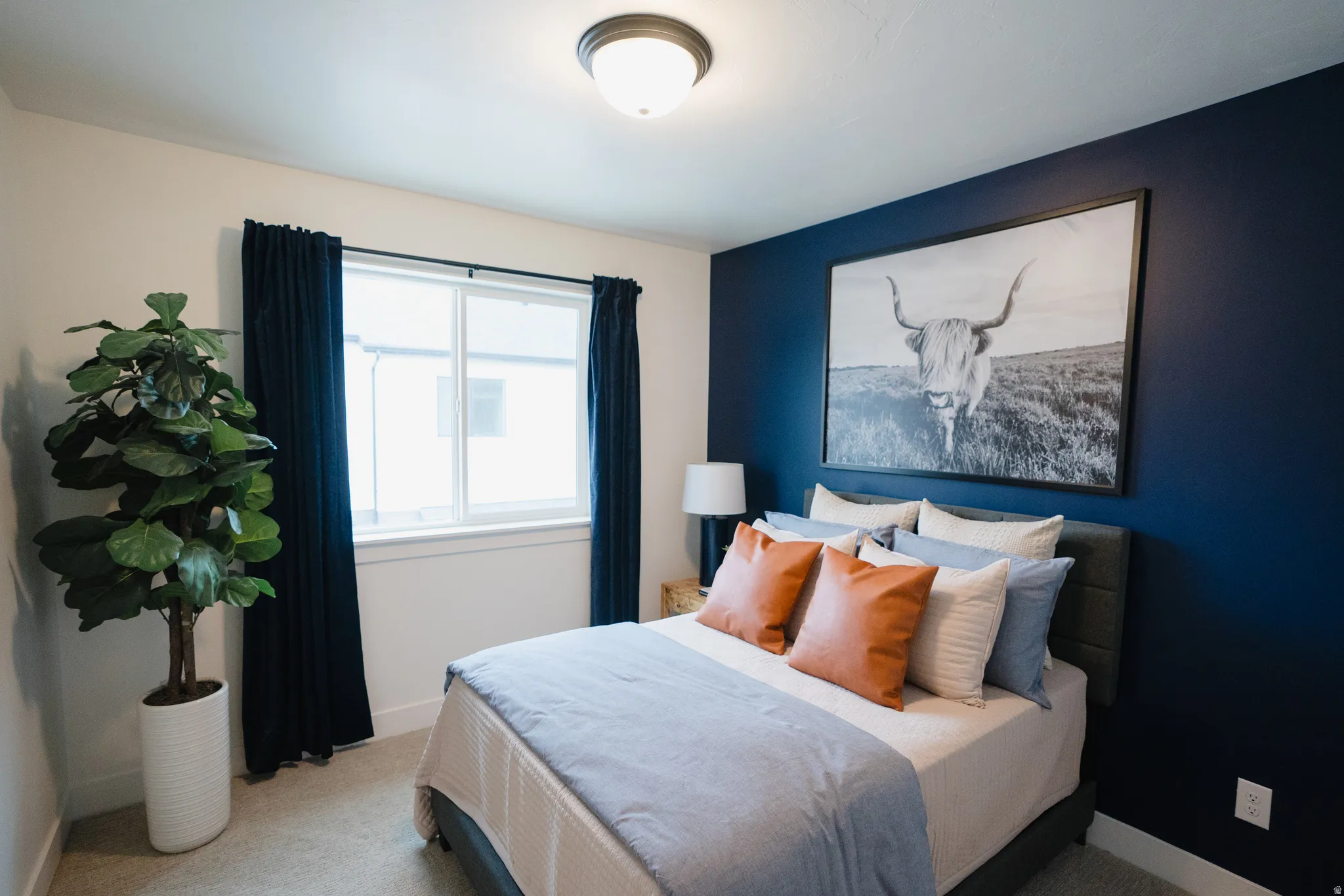 Bedroom featuring a window with dark curtains, a deep blue accent wall, a flush-mount ceiling light fixture, and light-colored carpeting