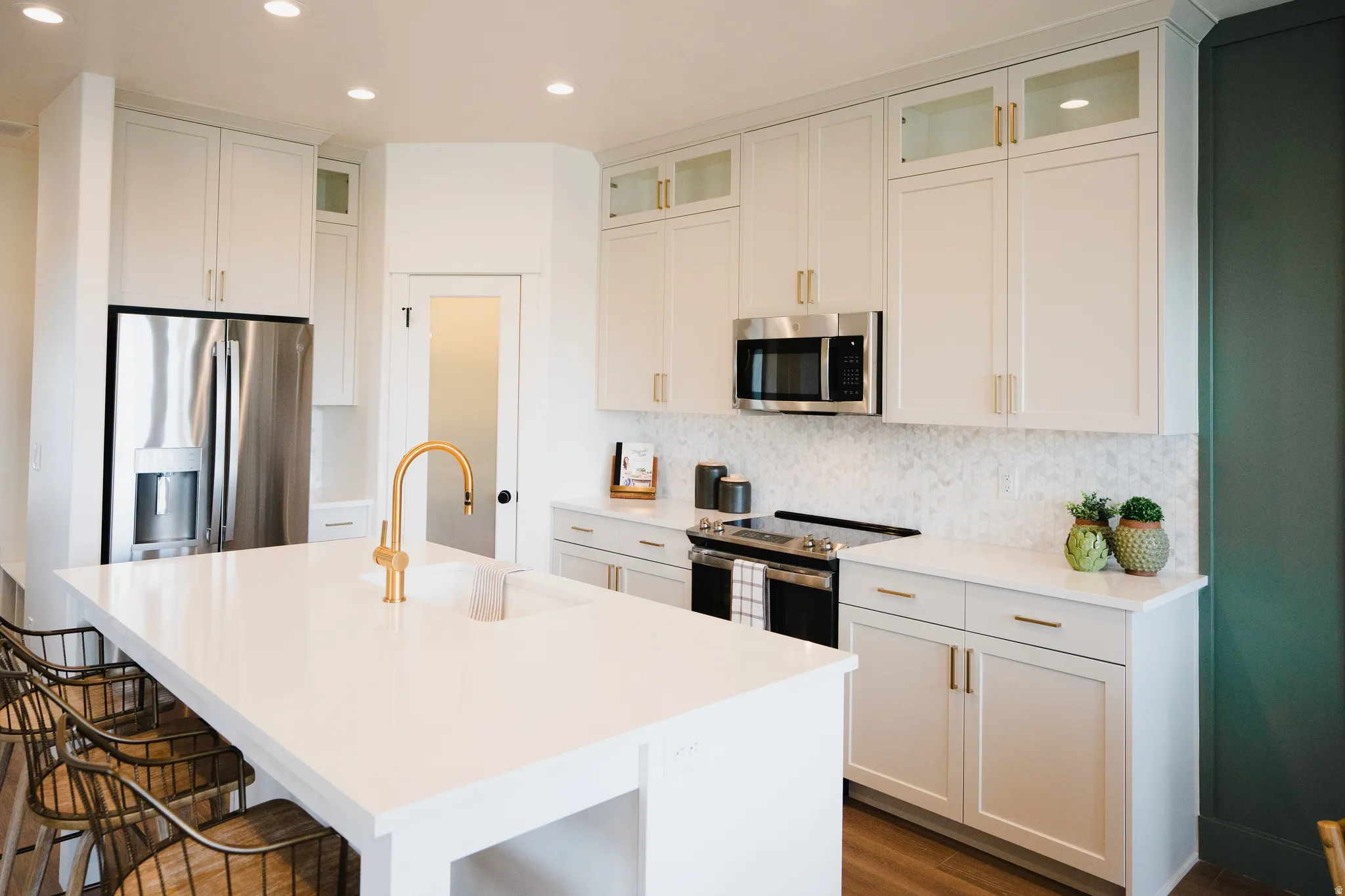 Contemporary kitchen featuring an expansive island with an integrated sink and gold-tone faucet, white shaker cabinetry with gold-tone hardware, stainless steel appliances, and a marble-finish backsplash