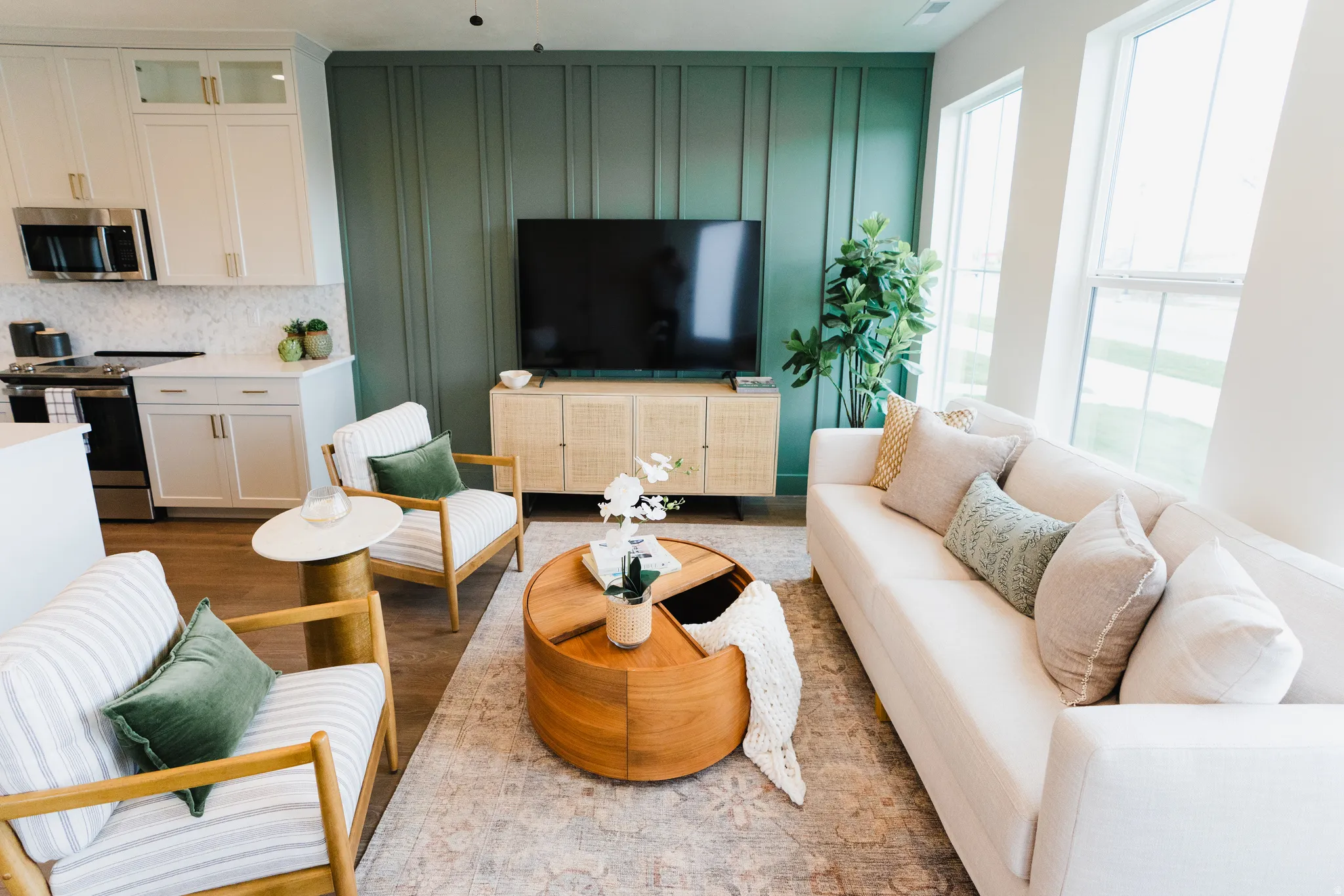 Open-concept living area featuring a striking paneled accent wall, wood-finish flooring, abundant natural light from large windows, a white kitchen with shaker cabinetry, and stainless steel appliances