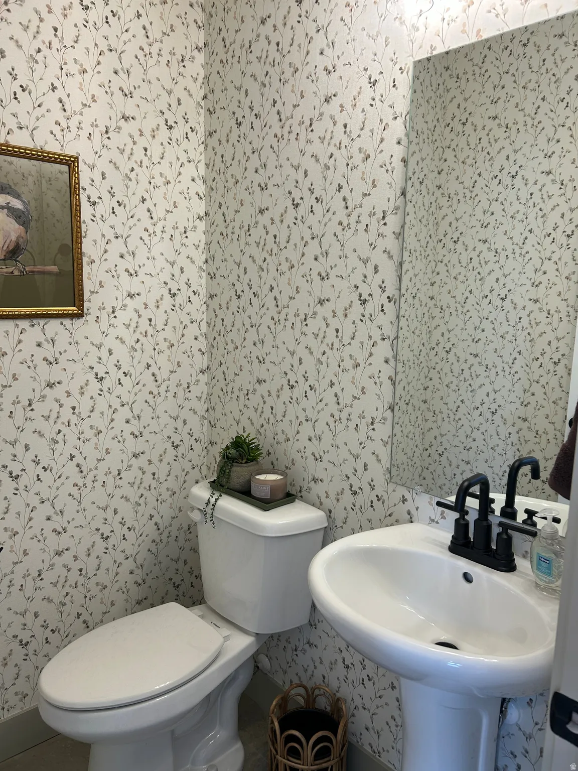 Pedestal sink with matte black faucet, wall-mounted mirror, and botanical print wallpaper