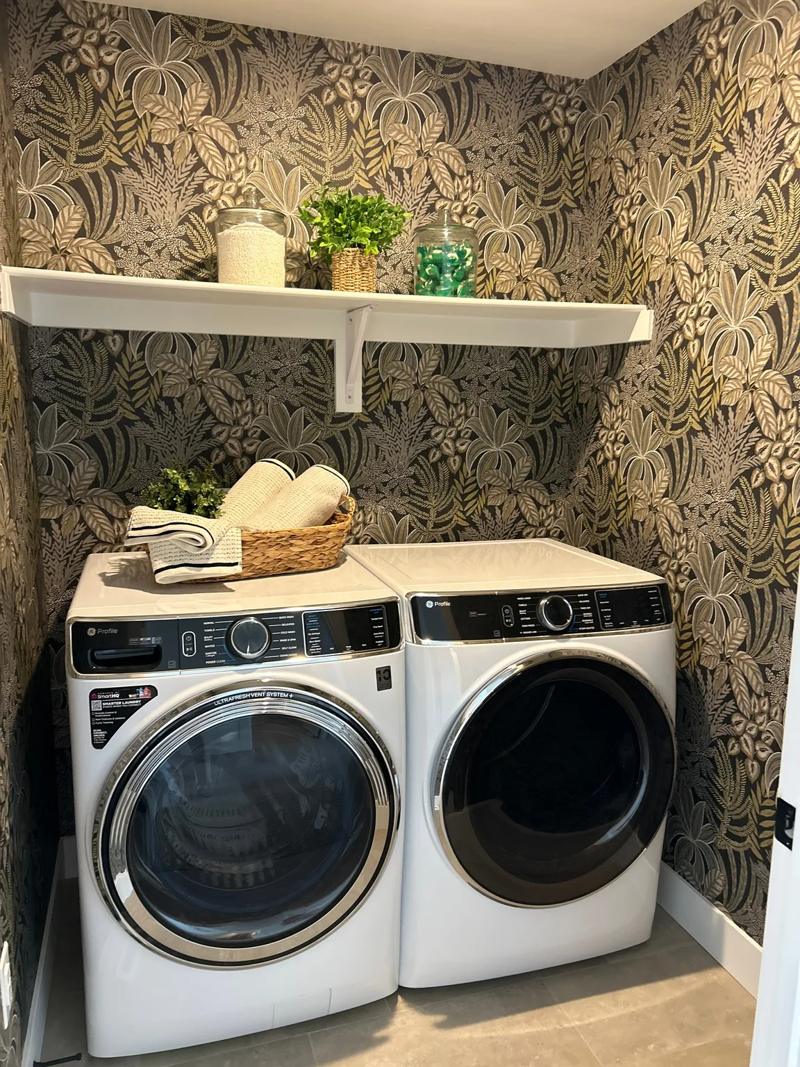 Dedicated laundry space featuring a white wall-mounted shelf, contemporary botanical wallpaper, and durable tile flooring