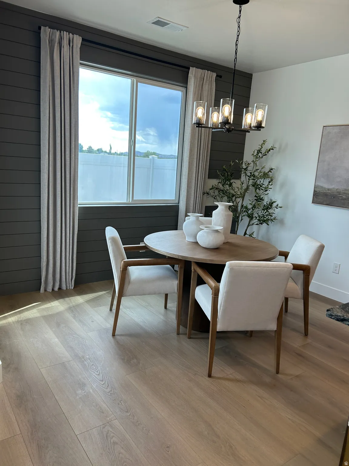Dining area featuring a modern chandelier, a large window, horizontal wood-panel accent wall, light wood-finish flooring, and light-colored drapery panels