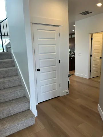Light wood-finish flooring extending from the hallway, a paneled white door with matte black hardware, and an adjacent carpeted staircase with a black metal handrail