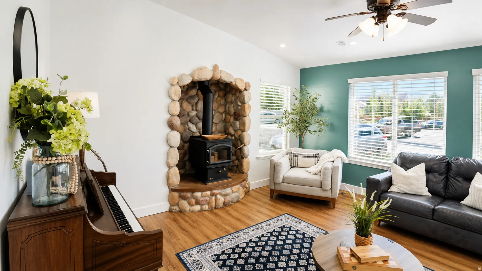 Living area featuring a stone wood-burning stove with a hearth, wood-finish flooring, a ceiling fan, and multiple windows with blinds