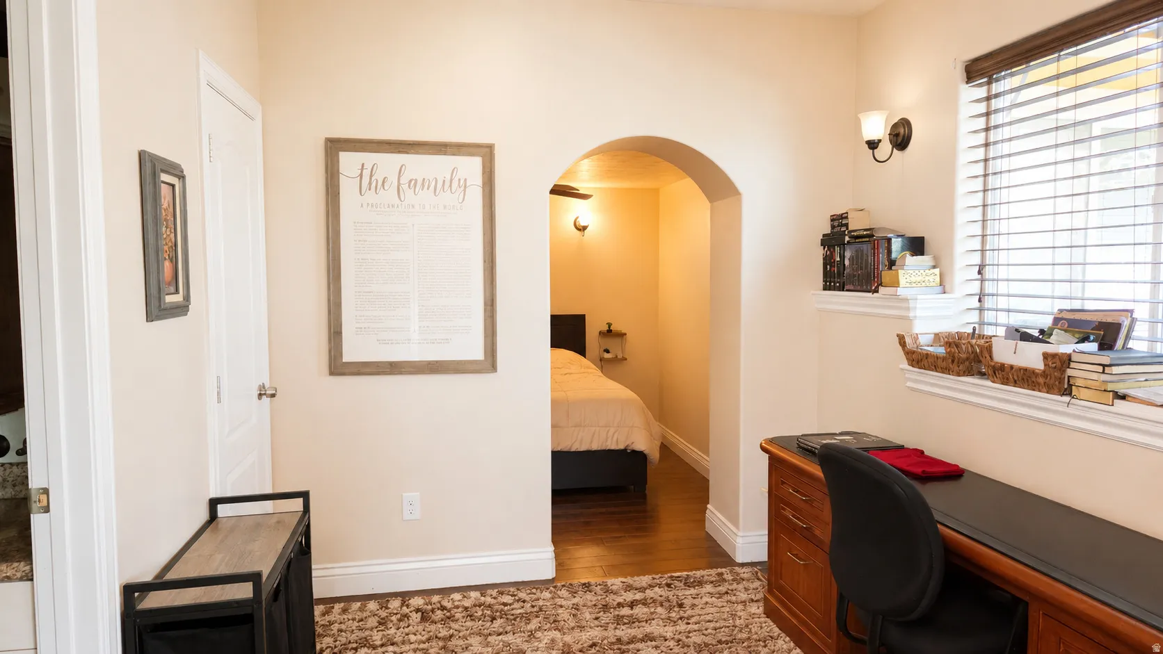 ADU #1 - Archway leading to a bedroom with wood-finish flooring, featuring a window with horizontal blinds and a wall-mounted sconce